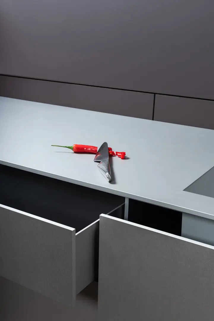 Eine in Scheiben geschnittene rote Chilischote und ein Metallmesser liegen auf einer hellgrauen Küchenarbeitsplatte aus Aluminium, darunter zwei offene Schubladen. Im Hintergrund sind minimalistische, moderne Schränke in dunklen Grautönen zu sehen.