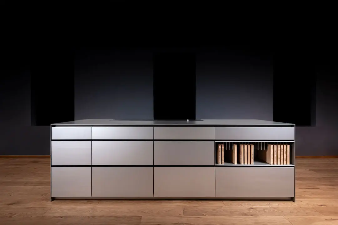Diese Künchendesign-Fotografie einer moderne, minimalistische Kücheninsel aus Aluminium mit schlichten grauen Schubladen und einem offenen Regal mit ordentlich angeordneten Büchern auf einem Holzboden vor einem dunklen Hintergrund.