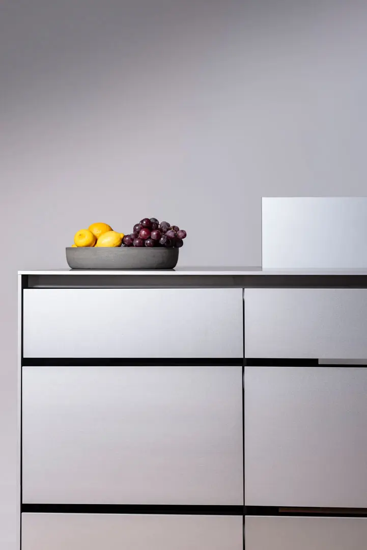Eine minimalistische Arbeitsplatte aus Aluminium ist Teil einer Küche des Designers Jan-Erik Heym mit einer grauen Schale mit gelben Äpfeln und roten Trauben, die auf modernen, hellgrauen Schubladen vor einer schlichten hellgrauen Wand steht.