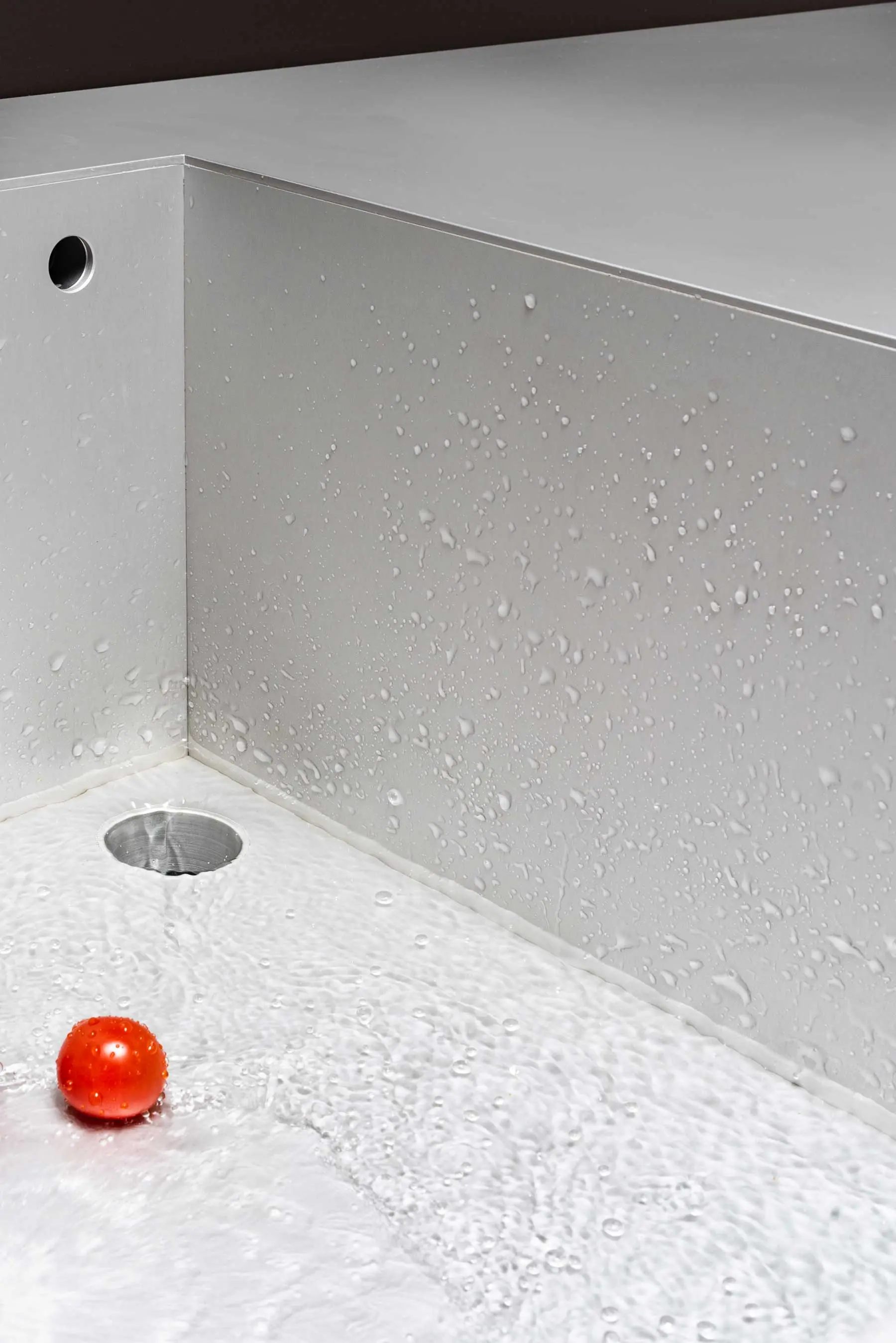 Eine rote Kirschtomate sitzt in der Nähe eines Abflusses in einem sauberen, Waschbecken aus Aluminium mit Wassertropfen auf der hellgrauen Oberfläche und den Wänden. Das Wasser fließt sanft über den Boden des Waschbeckens.