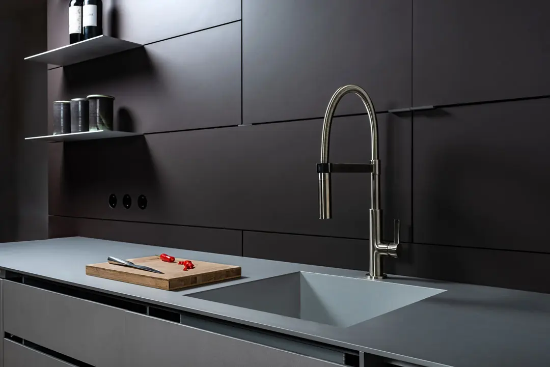 Eine moderne Küche aus Aluminium mit dunklen Schränken, einem eleganten Metallhahn, einer eingebauten Spüle, einem hölzernen Schneidebrett mit geschnittenen roten Chilischoten und Regalen mit Gläsern und Flaschen vor einer matten Wand.
