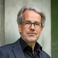 Holger Hafner trägt eine dunkle Jacke und ein schwarzes Hemd, steht vor einem neutralen Hintergrund und blickt mit einem leichten Lächeln in die Kamera.