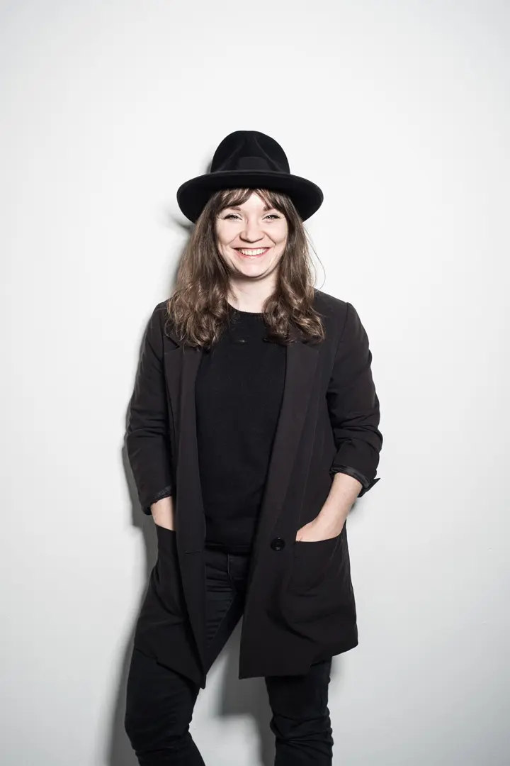 Lucia Bartl mit langen braunen Haaren, die einen schwarzen Hut, einen schwarzen Blazer und eine schwarze Hose trägt, steht vor einem schlichten weißen Hintergrund und lächelt mit den Händen in den Taschen.