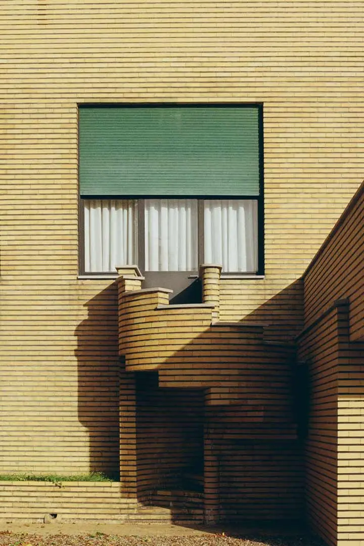 Die Villa Cavrois, ein gelbes Backsteingebäude mit einem großen grünen Fensterschirm, weißen Vorhängen und einer kleinen Wendeltreppe, die zu einer Tür führt. Die Schatten, die von der Treppe und dem Geländer geworfen werden, erzeugen geometrische Muster an der Wand.
