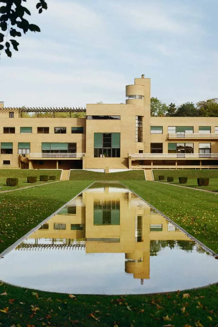Ein modernes, hellbraunes Backsteingebäude im Stil von Art-Déco Architektur mit grünen Fenstern spiegelt sich in einem langen, rechteckigen Pool, umgeben von gepflegten Rasenflächen, Hecken und verstreutem Herbstlaub unter einem klaren Himmel.