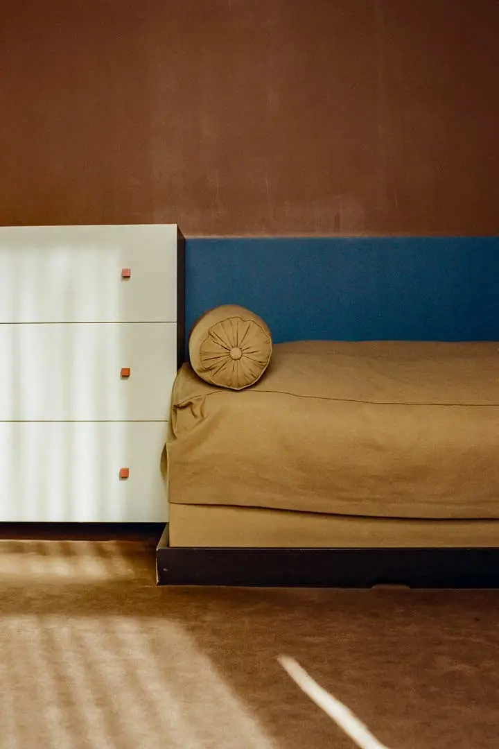 Ein minimalistisches Schlafzimmer mit einem hellbraunen Bett, einem zylindrischen Kissen, einem blauen Kopfteil und einer weißen Kommode mit braunen Knöpfen. Das Sonnenlicht wirft gestreifte Schatten auf den Boden und die Kommode. Die Wände sind braun gestrichen.