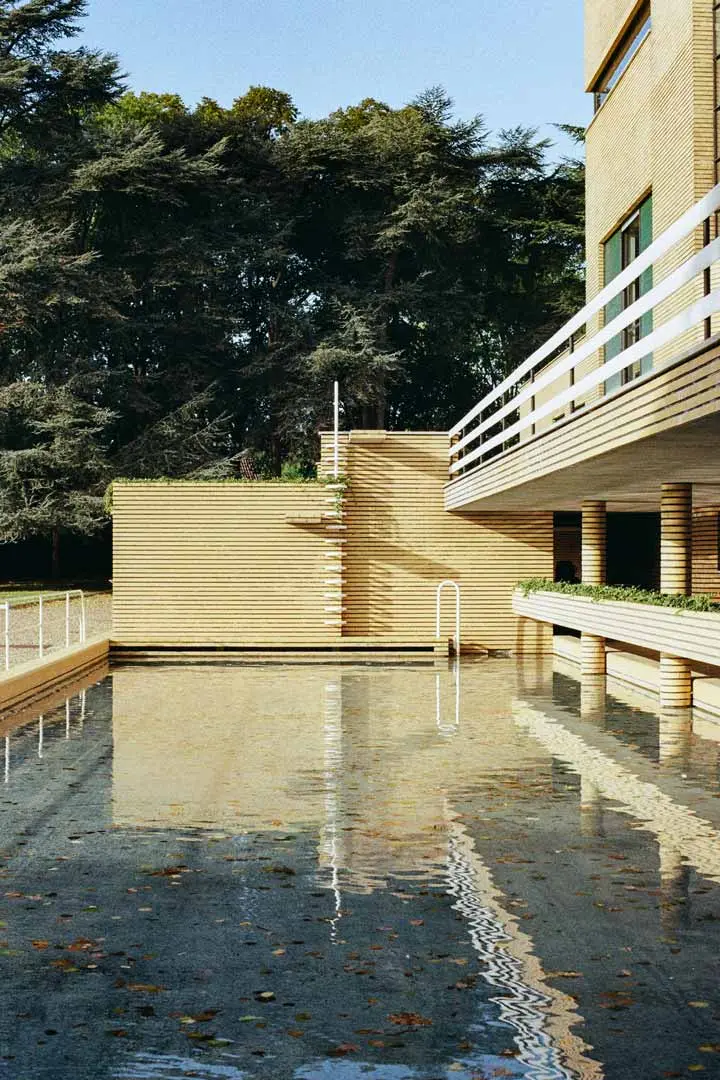 Ein modernes Gebäude im Stil der Art-Déco Architektur mit horizontalen Linien und einem Balkon blickt auf ein rechteckiges, spiegelndes Becken mit verstreuten Blättern. Im Hintergrund sind große Bäume und ein klarer blauer Himmel zu sehen.