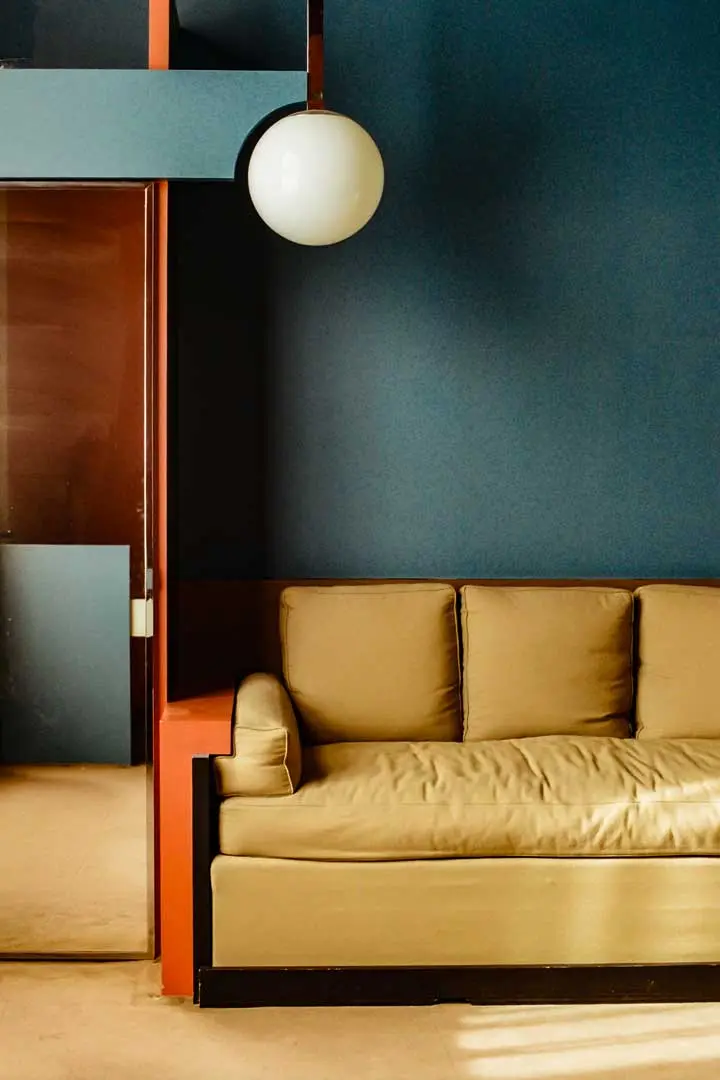 Ein modernes Wohnzimmer mit einem hellbraunen Sofa, blauen Wänden, roten architektonischen Akzenten, einer runden weißen Deckenleuchte und Sonnenlicht, das auf den Boden fällt.