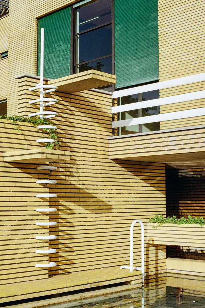 Ein modernes gelbes Backsteingebäude mit grünen Fenstern und einer einzigartigen weißen Wendeltreppe mit Geländer, einem kleinen Überhang mit Pflanzen und einem flachen Wasserbecken darunter.