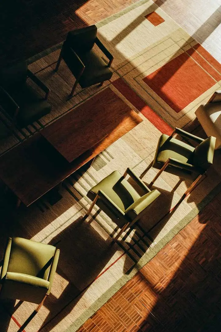 Aufnahme von oben von fünf grün gepolsterten Stühlen und einem Holztisch auf einem gemusterten Teppich, wobei das starke Sonnenlicht lange Schatten wirft - eine zeitlose Anmutung von analoger Architekturfotografie, die an das Projekt Villa Cavrois erinnert.