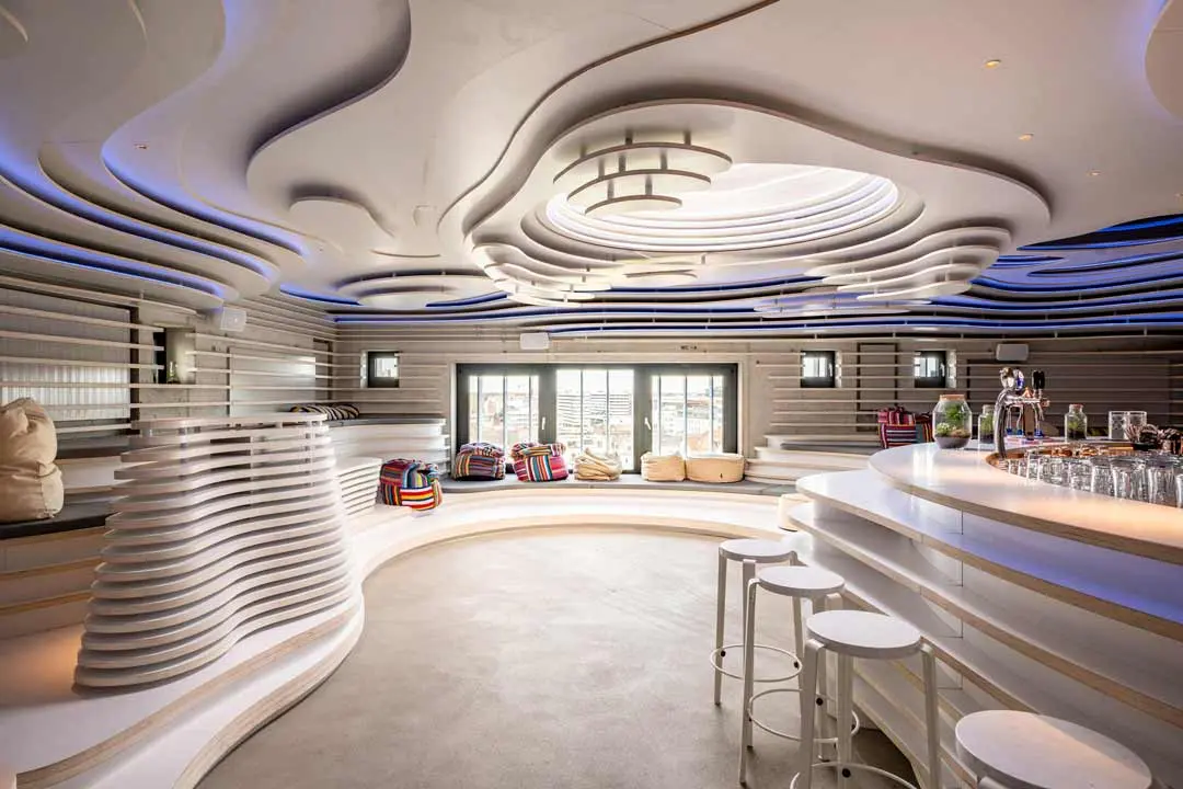 Roof Drop Bar der Villa Viva, eine moderne Lounge mit gewellten, geschichteten weißen Wänden und Decken, einem zentralen Oberlicht, einer geschwungenen Bar mit weißen Hockern und bunten Sitzsäcken an großen Fenstern. Der Raum hat ein futuristisches, minimalistisches Design.