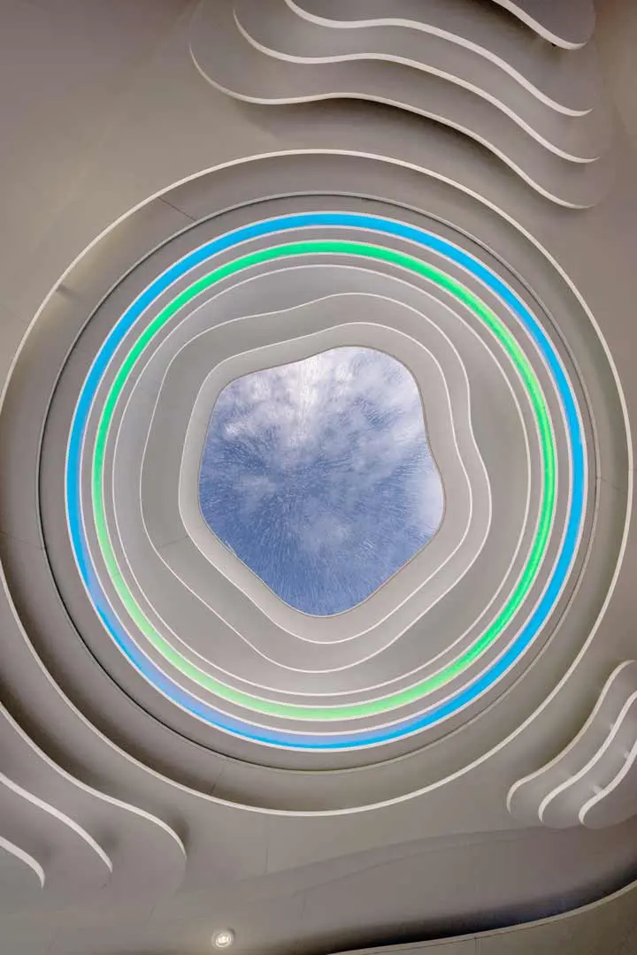 Ein rundes Oberlicht mit einem unregelmäßigen, gewellten Rand ist von konzentrischen Ringen aus sanften weißen, grünen und blauen Lichtern umgeben, über denen der Blick auf einen bewölkten Himmel schweift. Die Decke weist geschwungene, futuristische Muster auf. Fenster der Roofdrop Bar der Villa Viva, Hamburg.