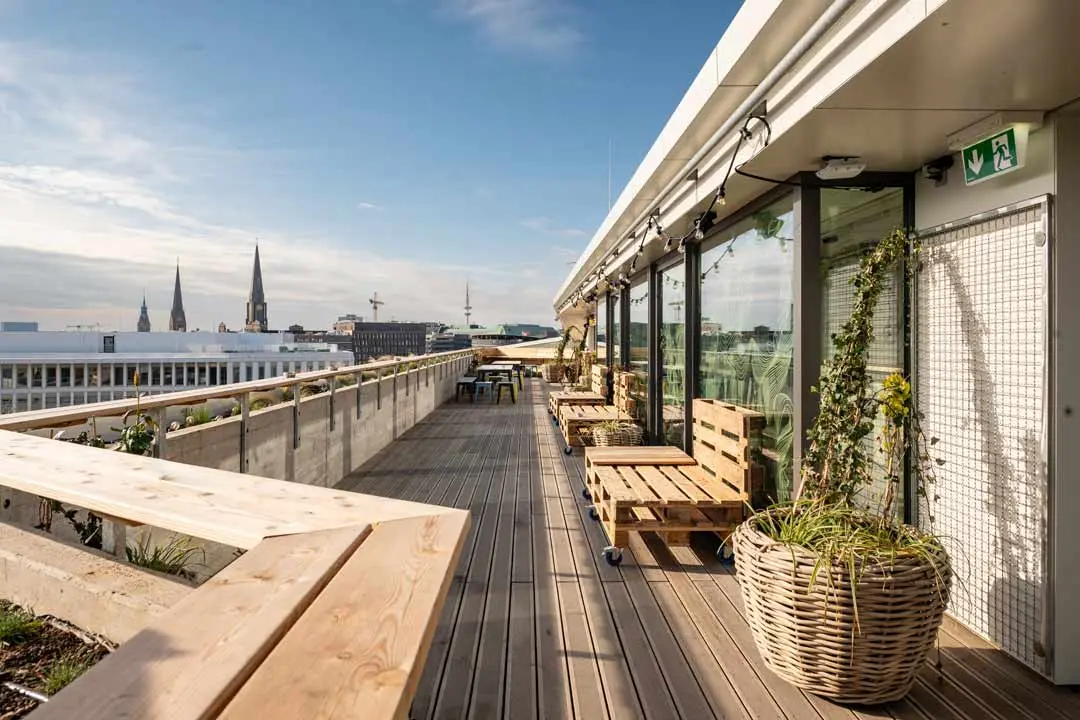 Geräumige Dachterrasse der Roof Drop Bar mit Holzbelag, Bänken und Topfpflanzen überblickt man die Skyline der Stadt mit Kirchtürmen in der Ferne unter blauem Himmel.