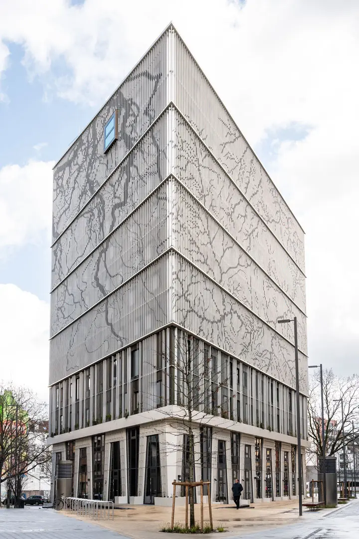 Das Hotel Villa Viva in Hamburg mit vertikalen Metalllamellen und verschlungenen, organischen Mustern an der oberen Fassade. Das Gebäude hat große Fenster in den unteren Stockwerken und ein einzelnes Fenster in der Nähe des Daches. Davor stehen ein Baum und eine Person.