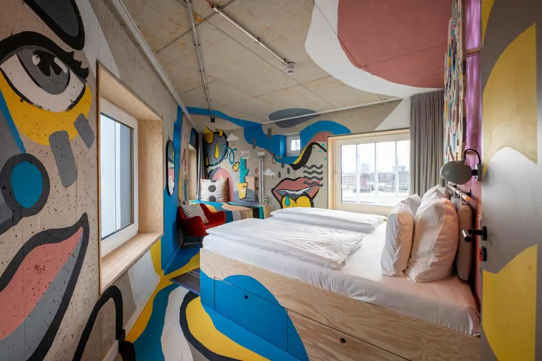 Ein Hotelzimmer, dessen Wände und Decke mit farbenfroher abstrakter Wandmalerei mit markanten Gesichtern und Formen bedeckt sind. Das Zimmer hat ein großes Bett, zwei Fenster, einen Schreibtisch und eine einzigartige künstlerische Einrichtung.