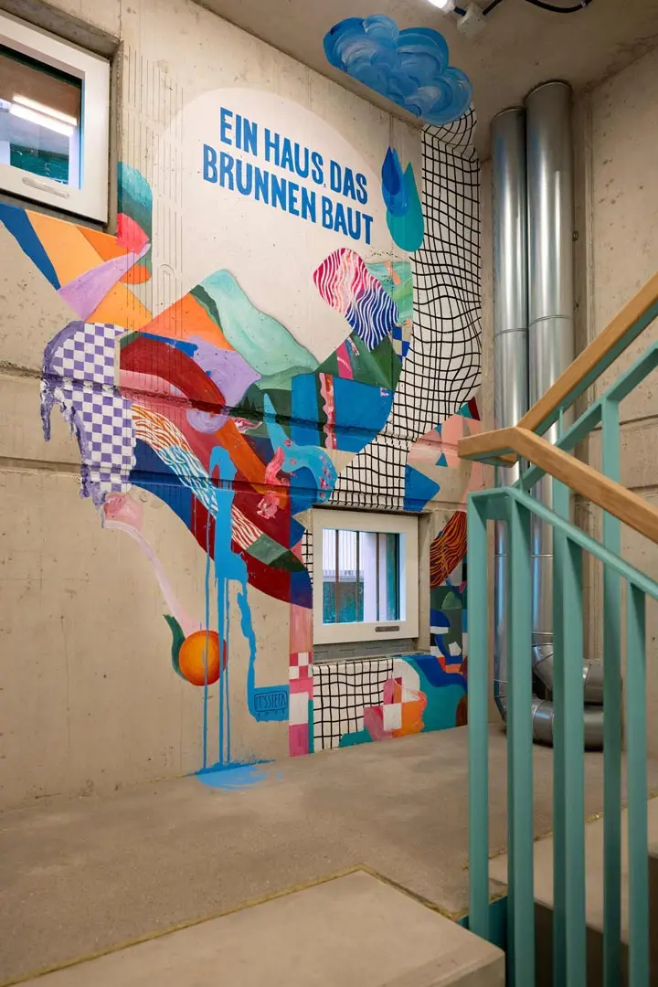 Treppenhaus der Villa Viva, Hamburg ist Teil der Serie Hotelfotogragie. Buntes abstraktes Wandbild mit geometrischen Formen und Mustern schmückt eine Betontreppenhauswand. Der blaue Text oben lautet EIN HAUS, DAS BRUNNEN BAUT. Ein hellgrünes Geländer und kleine Fenster sind sichtbar.