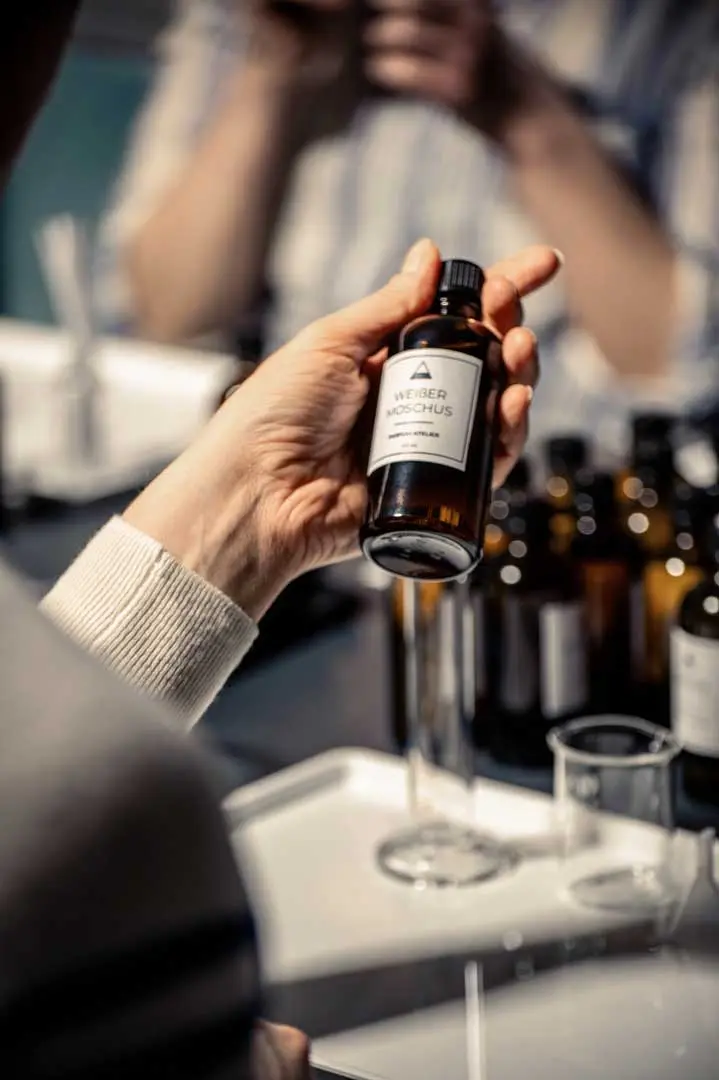 Eine Person hält eine kleine Braunglasflasche mit der Aufschrift WEIßER MOSCHUS über einen Tisch mit ähnlichen Flaschen, Glasbechern und weißen Tabletts, bei einem Parfum Workshop von Parfum Atelier, Hamburg.
