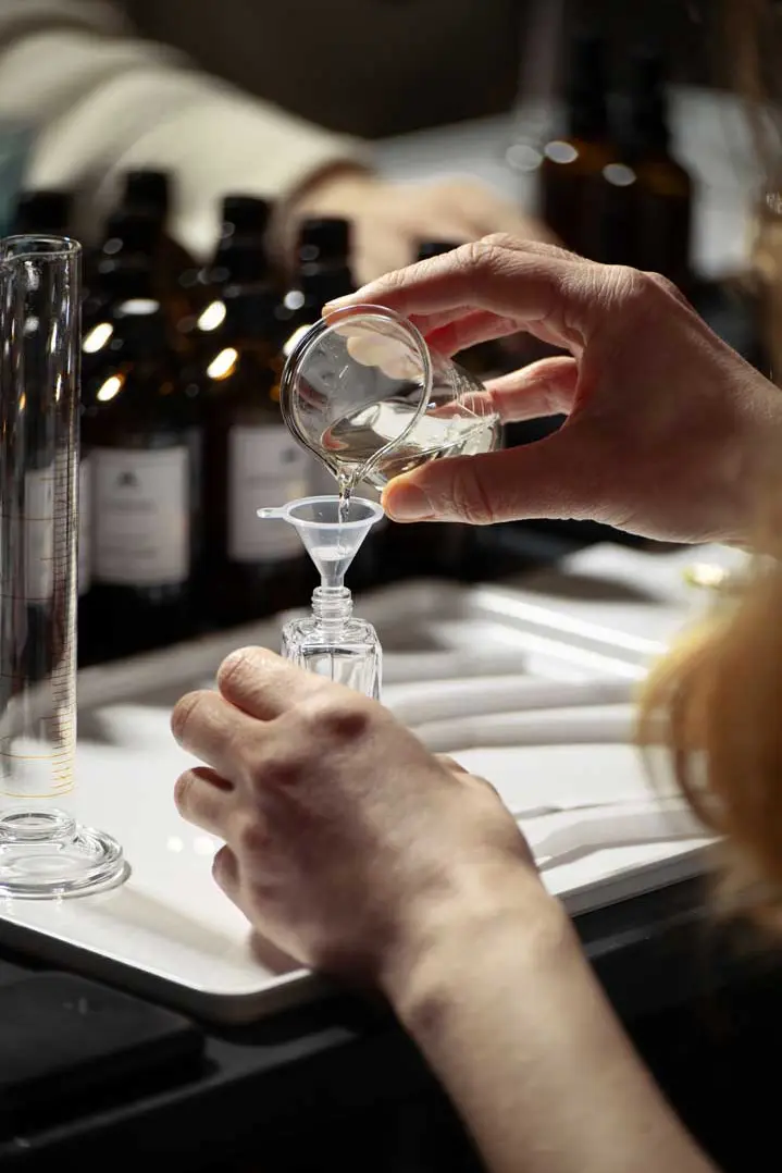 Eine Person gießt Parfum aus einem kleinen Glasbecher durch einen Trichter in eine Flasche, während im Hintergrund braune Glasflaschen und Messgeräte zu sehen sind. Das Bild ist Teil einer Social-Media Kampagne für das Unternehmen Parfum Atelier, Hamburg.