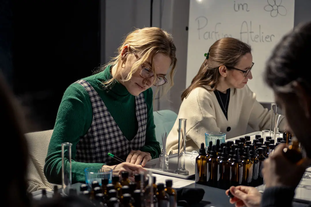 Zwei Frauen sitzen an einem Tisch, der mit braunen Glasflaschen und Laborgeräten aus Glas bedeckt ist, und schreiben konzentriert Notizen während eines Workshops zur Parfümherstellung. Auf einem Whiteboard im Hintergrund steht "Parfum Atelier".