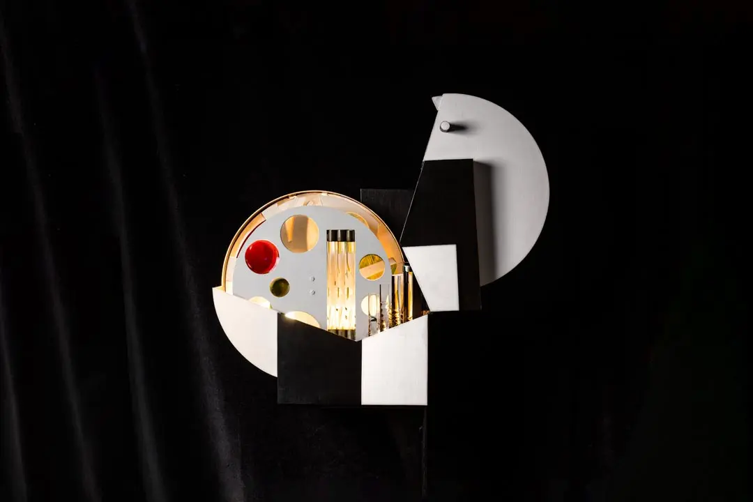 Eine geometrische, abstrakte Skulptur mit schwarzen, weißen und goldenen Formen mit kreisförmigen und rechteckigen Ausschnitten, die vor einem dunklen Hintergrund beleuchtet werden. Das Design ähnelt sich überschneidenden Kästen und Scheiben.