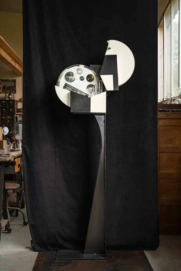 Eine hohe abstrakte Skulptur von Sylvain Larrière mit schwarzen und weißen geometrischen Formen, Kreisen und Spiegeln steht vor einem schwarzen Hintergrund in einer Künstlerwerkstatt, umgeben von Arbeitstischen und Schubladen.