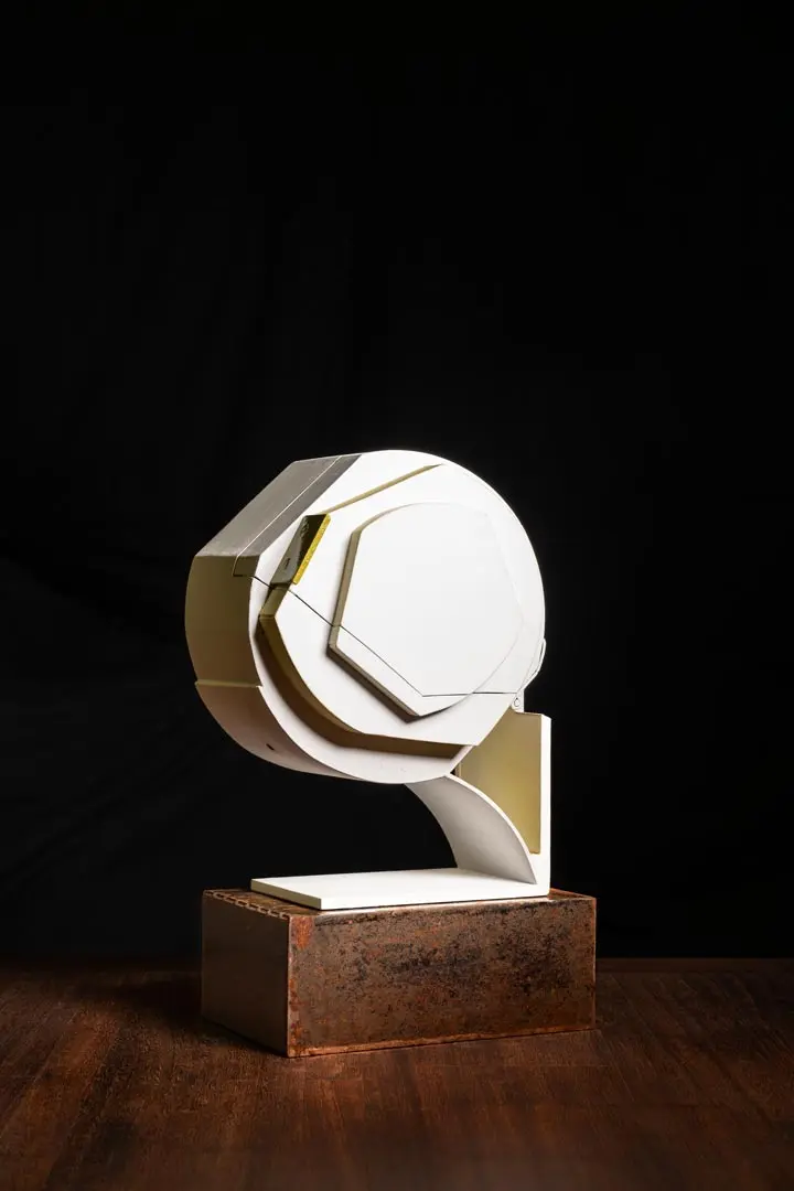 Eine moderne weiße, geometrische Skulptur mit eckigen und runden Formen steht auf einem braunen, rechteckigen Steinsockel, der vor einem schwarzen Hintergrund auf einer Holzfläche steht.