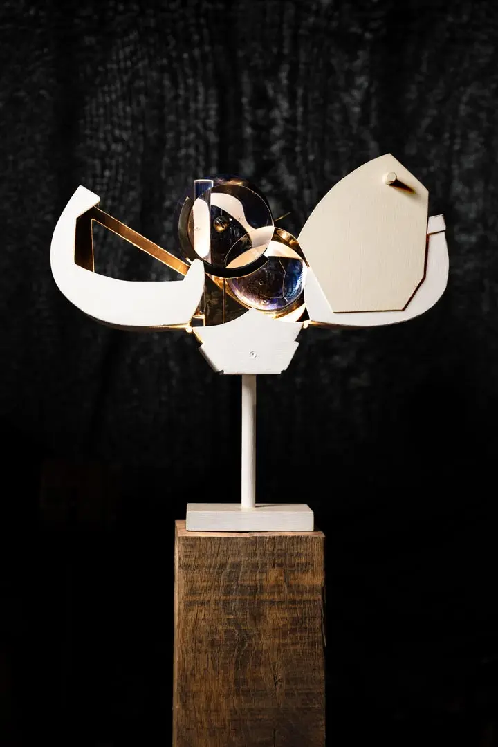 Eine moderne abstrakte Skulptur von Sylvain Larrière mit geometrischen weißen Formen und transparenten kreisförmigen Elementen, montiert auf einem weißen Ständer auf einem Holzblock, vor einem dunkel strukturierten Hintergrund.