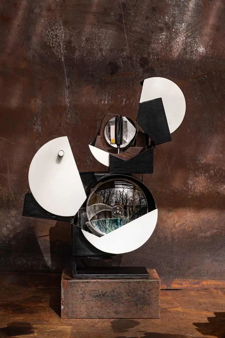 Abstrakte Metallskulptur mit schwarzen und weißen geometrischen Formen, darunter Kreise und Rechtecke, die auf einem rostfarbenen Hintergrund angeordnet sind. Das Kunstwerk von Sylvain Larrière reflektiert Licht und hat Glaselemente mit Naturmotiven im Inneren.