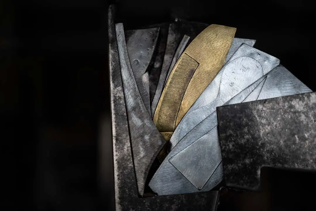 Nahaufnahme von geschichteten Metallteilen, darunter silber- und goldfarbene Bleche mit geometrischen Formen und scharfen Winkeln, die dicht vor einem dunklen Hintergrund gestapelt sind.
