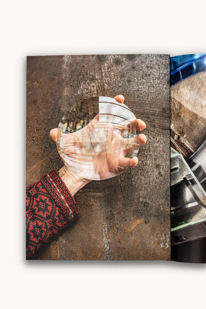 Katalog mit einer Hand vor einem Metallhintergrund hält eine große, ovale Glaslinse, die das Licht verzerrt und reflektiert und so ein verschwommenes Bild der Umgebung vor einem strukturierten braunen Hintergrund zeigt.