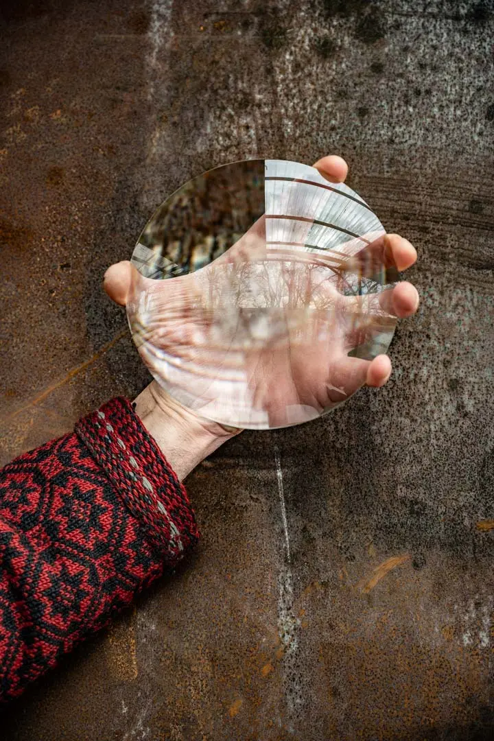 Eine Hand hält eine große, runde Lupe, das den Blick auf die Hand und den Hintergrund einer verrosteten, strukturierten Metalloberfläche verzerrt und vergrößert.