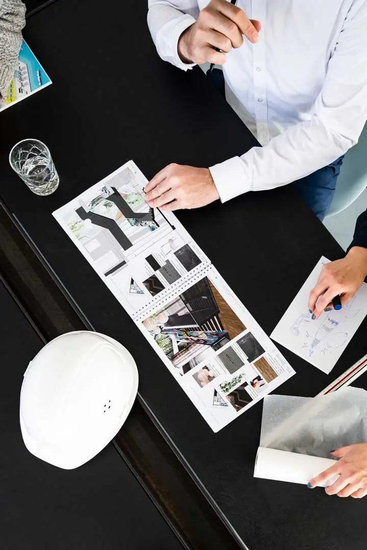 Das Foto zeigt zwei Personen, die an einem schwarzen Tisch sitzen und eine Broschür über Architektur- oder Designpläne sichten. In der Nähe sind gedruckte Bilder und Materialien, ein Wasserglas, ein weißer Schutzhelm und Skizzenblätter zu sehen.