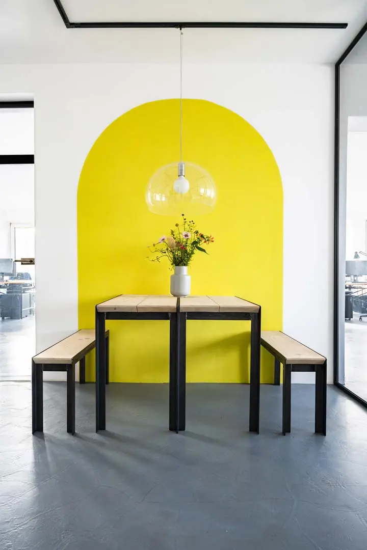 Ein kleiner Esstisch im Büro von Urban Future mit zwei Bänken steht an einer weißen Wand, auf die ein großer gelber Bogen gemalt ist. Das Foto zeigt eine Vase mit Blumen und eine runde Hängelampe befinden sich mittig über dem Tisch.