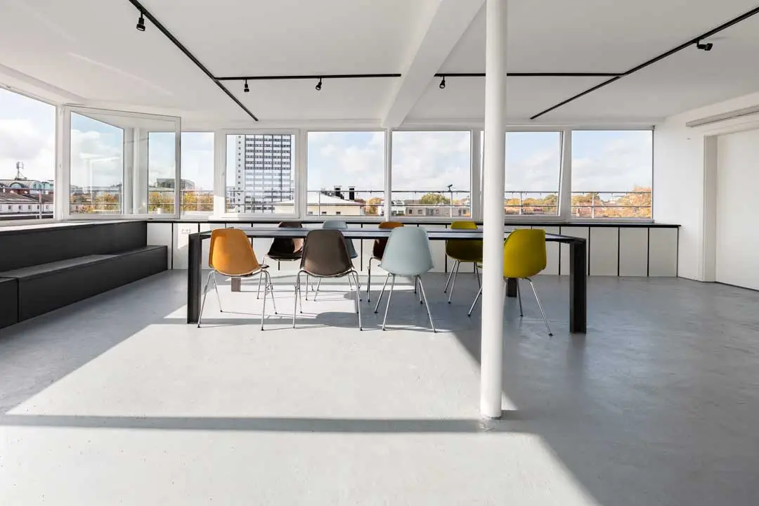 Ein moderner, minimalistischer Konferenzraum für Urban Future mit großen Fenstern, einem langen Tisch und bunten Stühlen. Das Sonnenlicht erfüllt den Raum, und von draußen kann man die Stadtlandschaft sehen. Der Raum hat einen hellen Boden und eine eingebaute Bank an einer Wand.