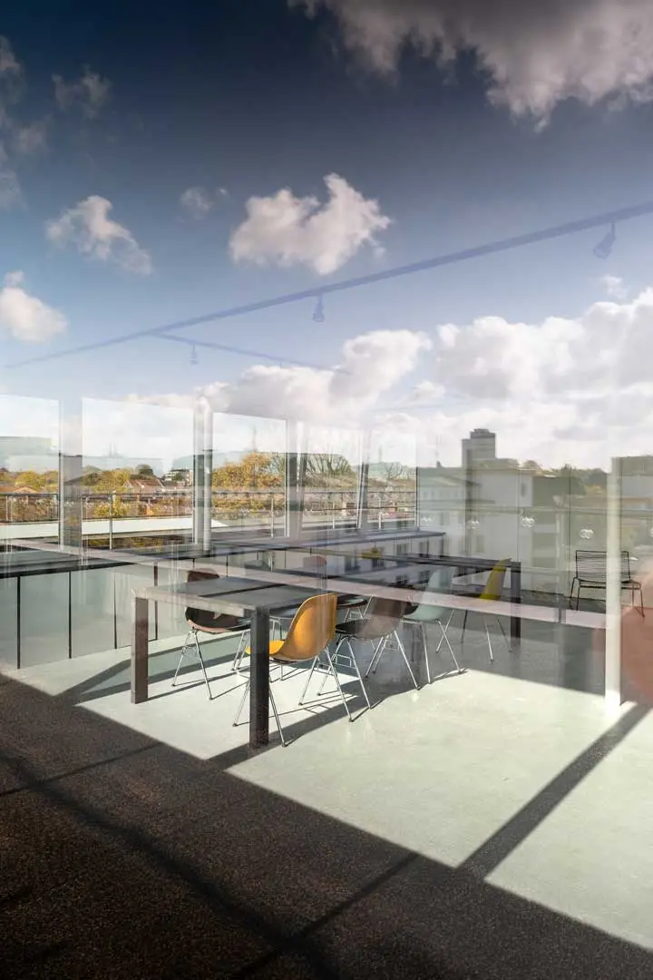 Moderner, sonnenbeschienener Konferenzraum mit Glaswänden und mehreren Stühlen um rechteckige Tische. Die Spiegelungen im Glas zeigen einen blauen Himmel mit Wolken und eine Stadtlandschaft im Hintergrund.