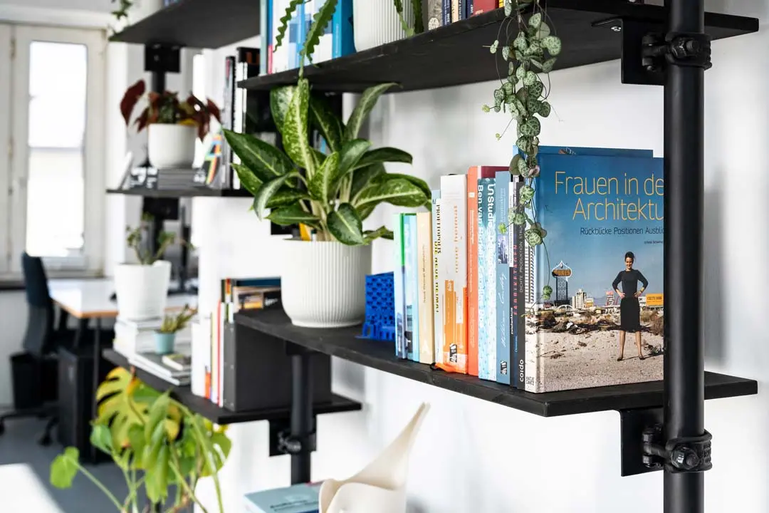 Corporate-Foto mit schwarzen, an der Wand befestigte Regal mit Büchern und mehreren grünen Topfpflanzen im Büro von Urban Future. Ein Buch mit dem Titel "Frauen in der Architektur" ist gut sichtbar nach außen gerichtet. Im Hintergrund ist ein Bürotisch zu sehen.
