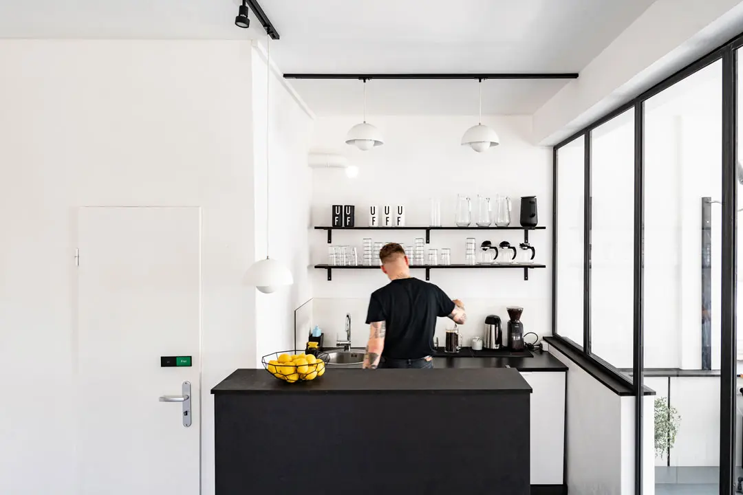 Corporate-Fotografie für Urban Future mit einem schwarz gekleideten Mann in einer modernen, minimalistischen Küche mit weißen Wänden und schwarzen Akzenten und bereitet ein Getränk zu. An der Wand befinden sich Regale mit Tassen und Gläsern, und auf dem Tresen steht eine Schale mit Zitronen.