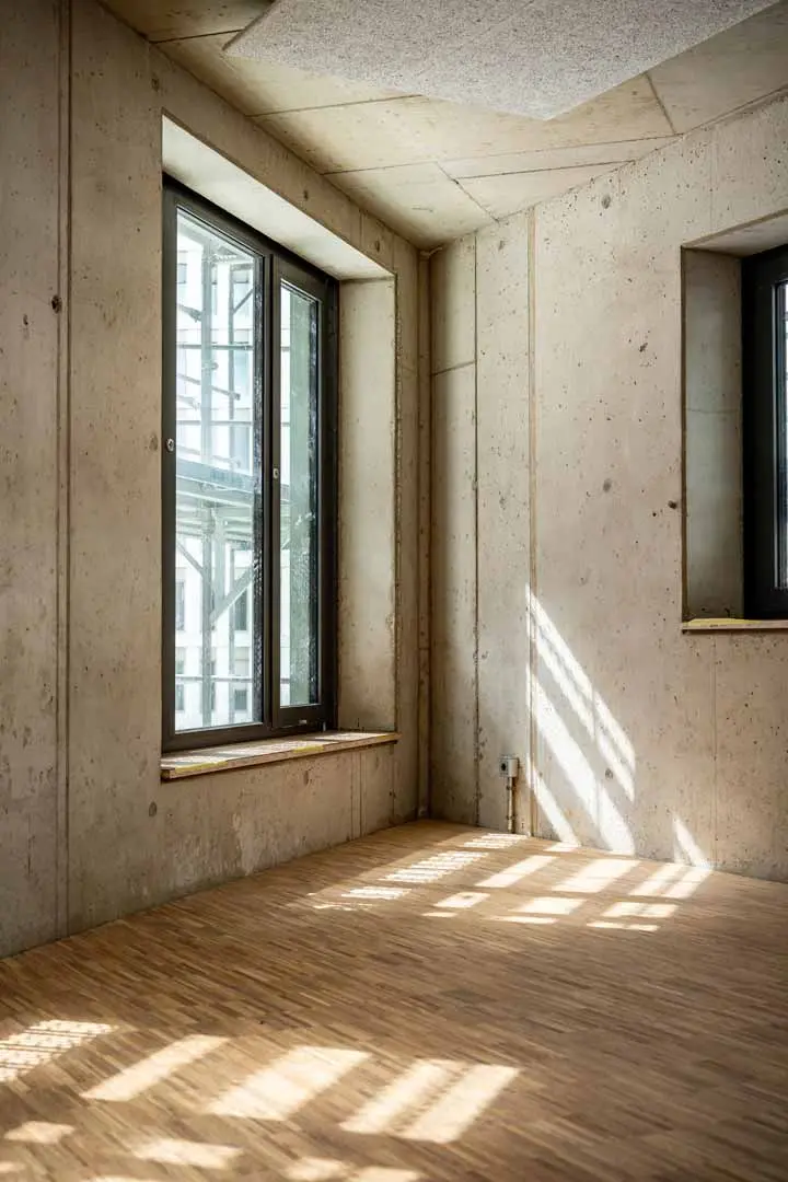 Das Sonnenlicht fällt durch große Fenster auf den Holzboden eines leeren Raums mit nackten Betonwänden und -decken und erzeugt geometrische Schatten auf den Oberflächen.