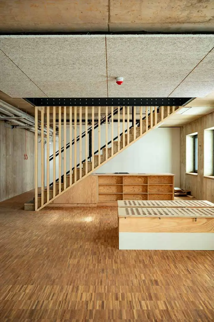 Ein modernes, sich im Bau befindliches Interieur mit Holzfußboden, einer offenen Treppe mit vertikalen Holzlatten, einer eingebauten Küchenzeile unter der Treppe und großen Fenstern an der rechten Wand, die natürliches Licht hereinlassen.