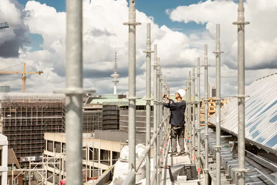 Ein Bauarbeiter mit Helm steht auf einem Baugerüst auf einem Gebäude und richtet eine Metallstange aus. Im Hintergrund sind der hamburger Fernsehturm und die Dächer der Innenstadt von Hamburg, Kräne und ein bewölkter Himmel zu sehen.