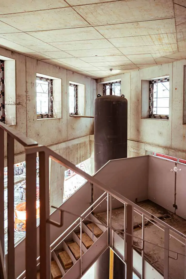 Ein großer, industriell anmutender Betonraum mit unfertigen Wänden, mehreren Fenstern, einer Metalltreppe und einem hohen zylindrischen Tank in der Mitte. Durch die Fenster sind Baumaterialien zu sehen. Dieses Bild ist Teil der Baudokumentation der Villa Viva.