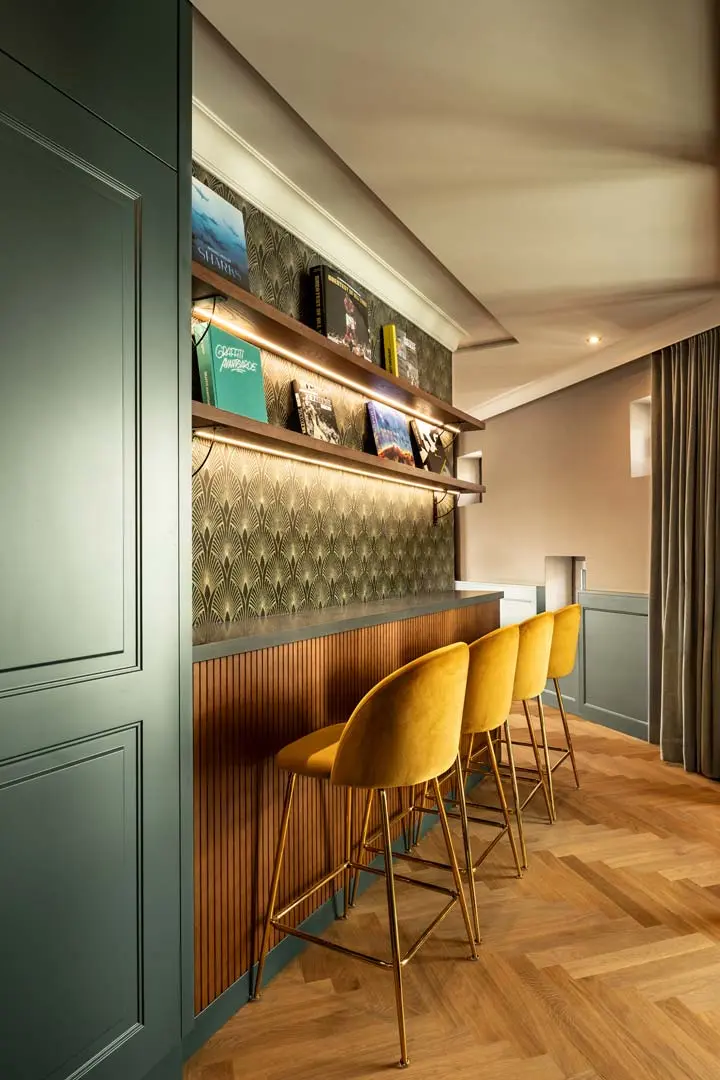 Eine stilvolle Hausbar mit drei gelben Barhockern aus Samt, gemusterter Aufkantung, Holzboden im Chevron-Design, Wandregalen mit Büchern und stimmungsvoller Beleuchtung. Der Raum ist modern eingerichtet und hat gedämpfte grüne Schränke.
