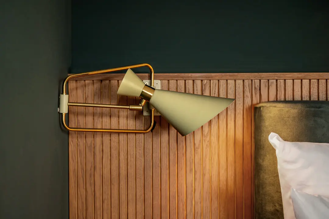 Eine moderne, gold- und beigefarbene Wandlampe mit geometrischem Muster ist an einem hölzernen Kopfteil mit Lattenrost neben einem Bett mit weißem Kissen vor einer dunkelgrünen Wand angebracht.