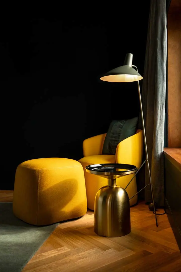 Diese gemütliche Leseecke mit einem gelben Sessel, einer passenden Ottomane, einem kleinen runden goldenen Beistelltisch, einer Stehlampe und einem grauen Vorhang steht vor einer dunklen Wand und einem Holzboden in der Jan Delay Suite.