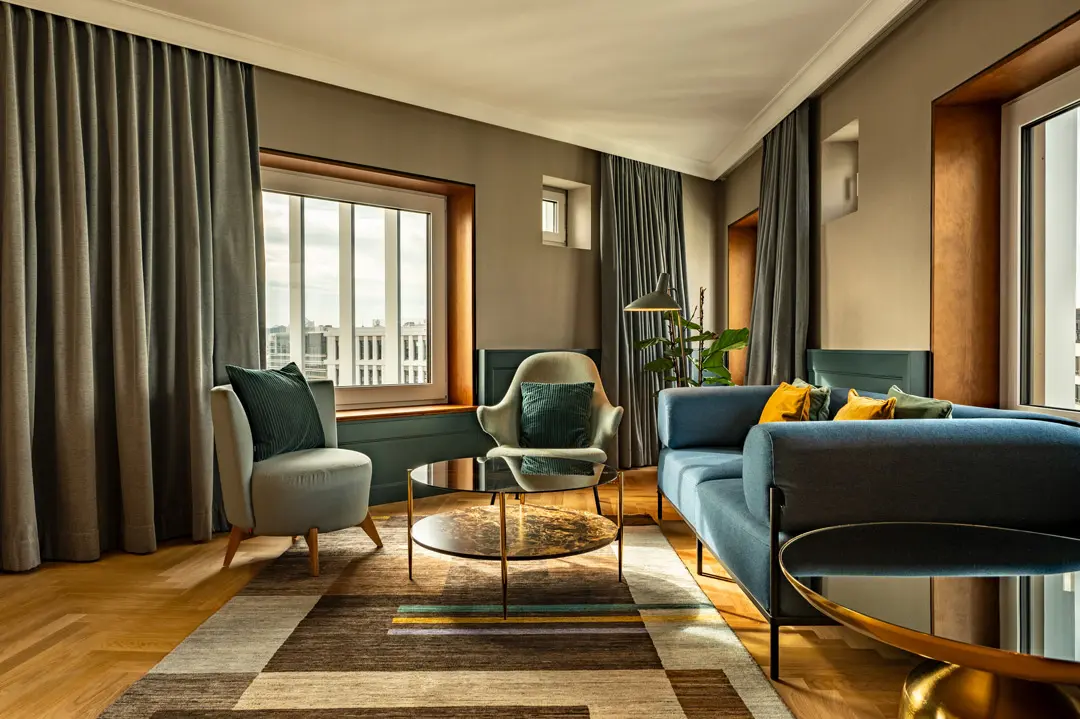 Dieses Hopitality-Foto zeigt ein modernes Wohnzimmer mit großen Fenstern, grauen Vorhängen, blauem Sofa, Sesseln, goldenen und gläsernen Couchtischen, einem Teppich mit geometrischen Mustern und einer Pflanze in der Ecke, wobei das natürliche Licht den Raum erhellt. Es ist ein Raum der Jan Delay Suite des Hotels Villa Viva.