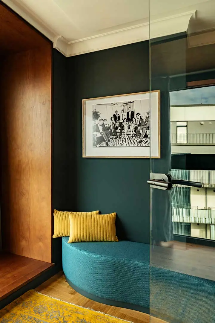 Eine gemütliche moderne Sitzecke mit einer geschwungenen tealfarbenen Bank, zwei gelben Kissen und einem gelben Teppich ist Teil der Jan Delay Suite im Hotel Villa Viva. Ein Schwarz-Weiß-Foto in einem Holzrahmen hängt an einer dunklen Wand. Glastür und Holzakzent sichtbar.
