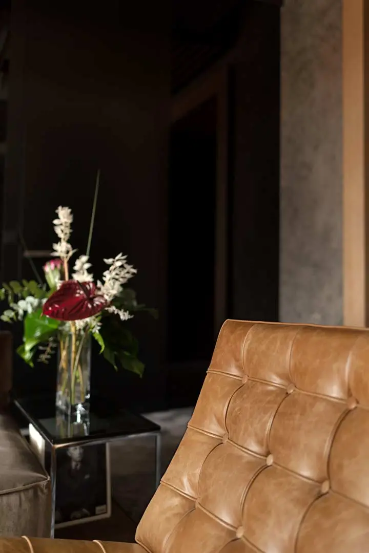 Eine Nahaufnahme eines hellbraunen, getufteten Ledersessels mit einer Glasvase mit verschiedenen Blumen auf einem Tisch im Hintergrund in einem sanft beleuchteten modernen Wohnzimmer.