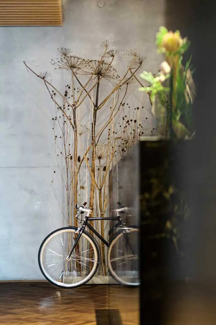 Die Fotografie aus der Serie Office Wunschhaus zeigt ein schwarzes Fahrrad, das in einem Innenraum vor einer grauen Wand steht, die mit hohen, getrockneten Pflanzen dekoriert ist. Im Vordergrund ist ein Teil eines unscharfen Blumenarrangements zu sehen.