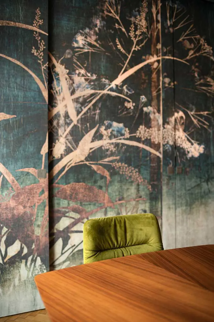 Ein grüner Samtstuhl steht an einem Holztisch, im Hintergrund ein großes, kunstvolles Wandgemälde mit Silhouetten von Laub und Zweigen in gedeckten Tönen.