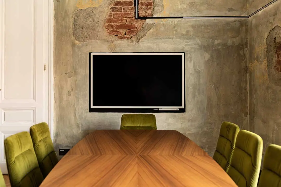 Ein moderner Besprechungsraum mit einem Holztisch, sechs grün gepolsterten Stühlen, einem an der Wand montierten Flachbildfernseher und verwitterten Sichtbetonwänden mit Ziegelsteinflecken.